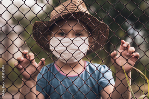 A child in a medical mask holds the fence with his hands. Quarantine melancholy and loneliness concepts © andrei310