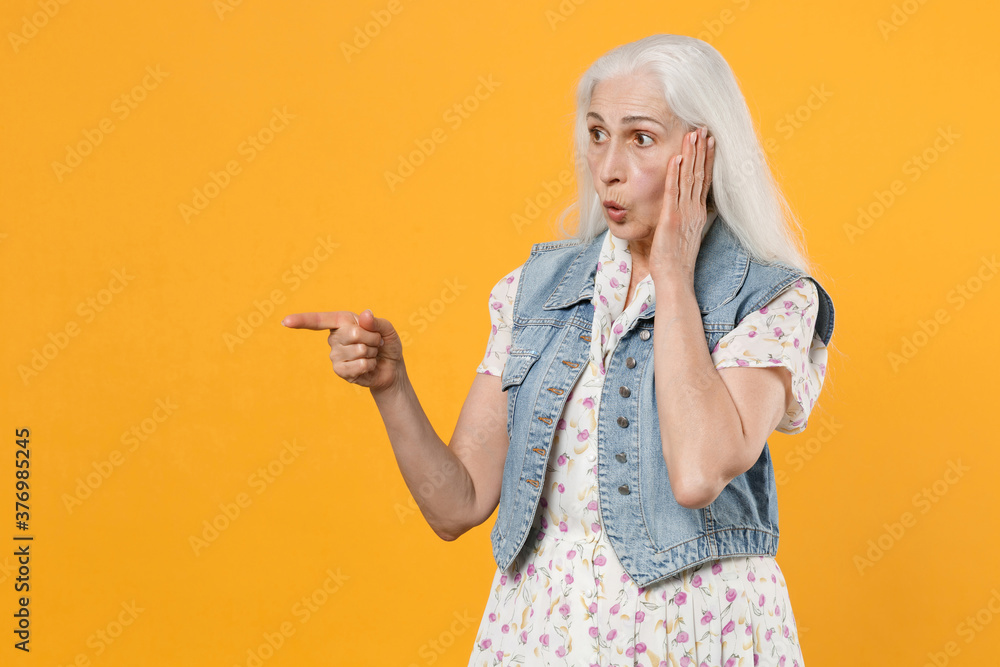 Amazed elderly gray-haired female woman in casual dress denim waistcoat posing pointing index finger aside on mock up copy space put hand on cheek isolated on yellow color background studio portrait. Amazed elderly gray-haired female woman in casual dress denim waistcoat posing pointing index finger aside on mock up copy space put hand on cheek isolated on yellow color background studio portrait.