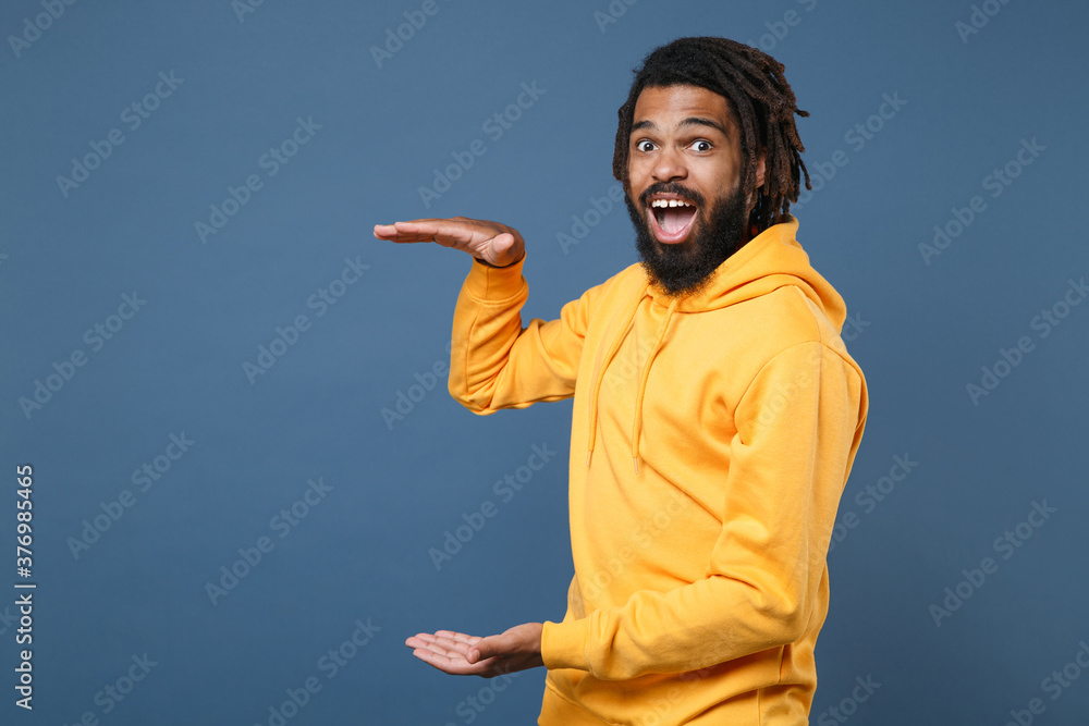 Naklejka premium Shocked young african american man guy in yellow streetwear hoodie isolated on blue wall background. People lifestyle concept. Mock up copy space. Gesturing demonstrating size with vertical workspace.