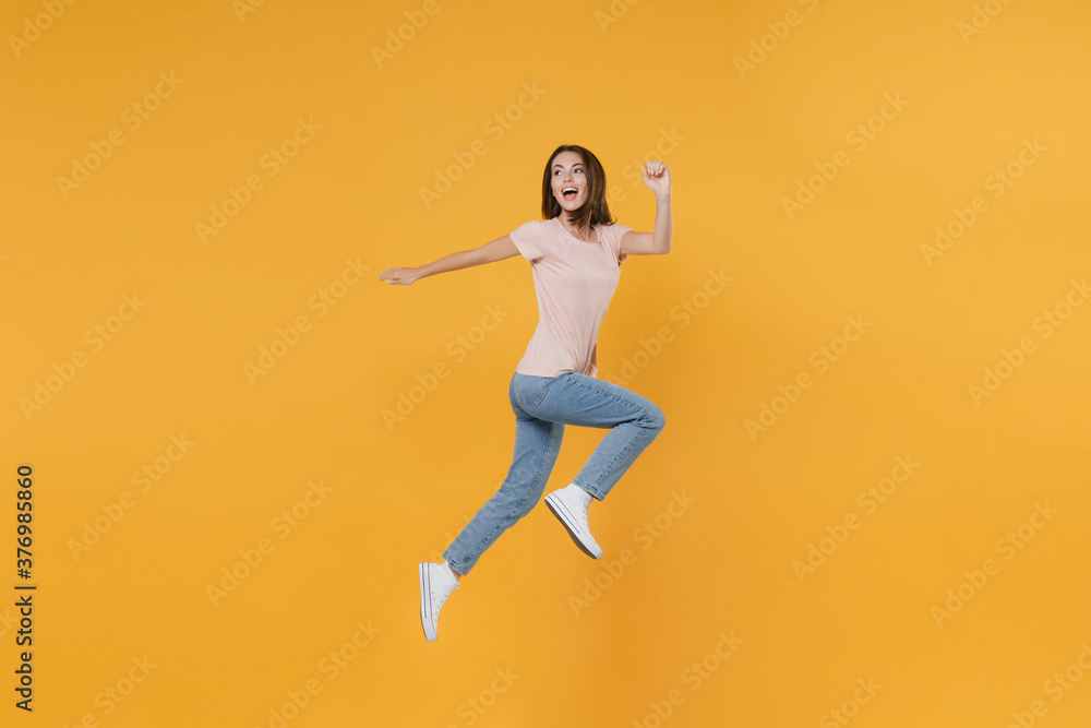 Full length side view portrait of cheerful funny young woman 20s wearing pastel pink casual t-shirt posing jumping like running looking aside isolated on bright yellow color wall background studio.