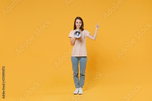 Full length portrait of smiling beautiful young woman wearing pastel pink casual t-shirt hold megaphone pointing index finger up on mock up copy space isolated on yellow color wall background studio. © ViDi Studio