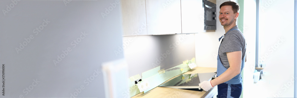 Portrait of cheerful handyman setting cookstove on kitchen. Professional service worker in uniform helping with moving. New property. Renovation and interior design concept Portrait of cheerful handyman setting cookstove on kitchen. Professional service worker in uniform helping with moving. New property. Renovation and interior design concept