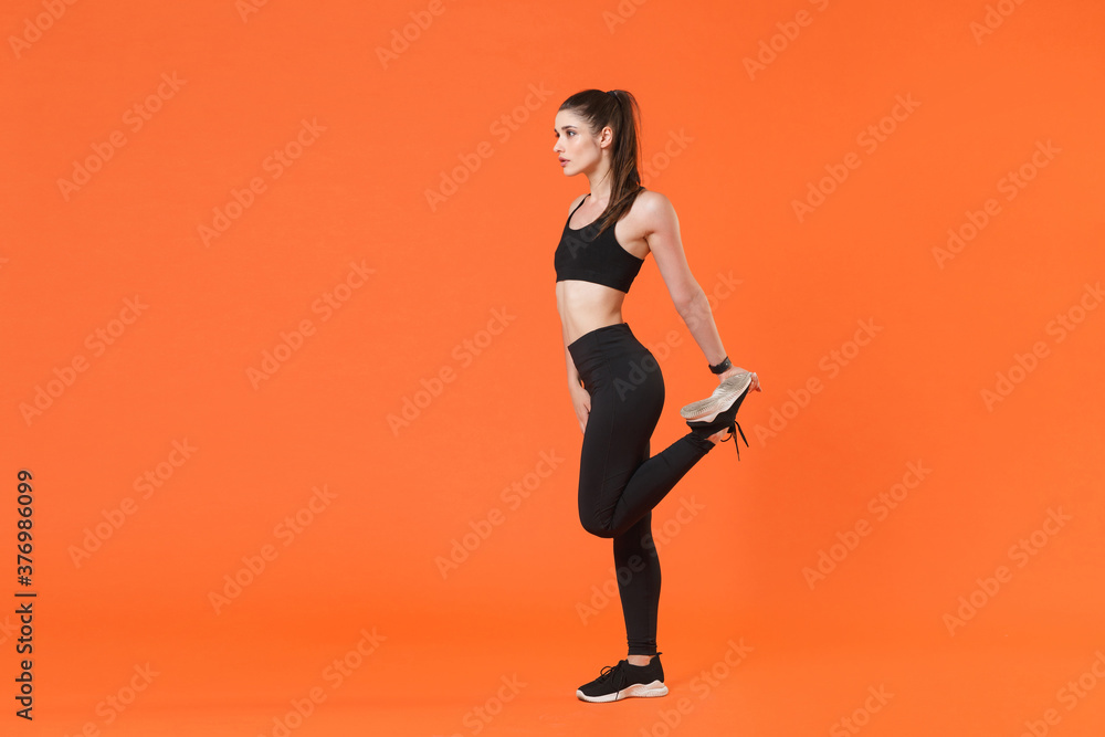 Full length side view portrait young fitness sporty woman in black sportswear posing training working out doing stretching exercising for legs looking aside isolated on orange color background studio.