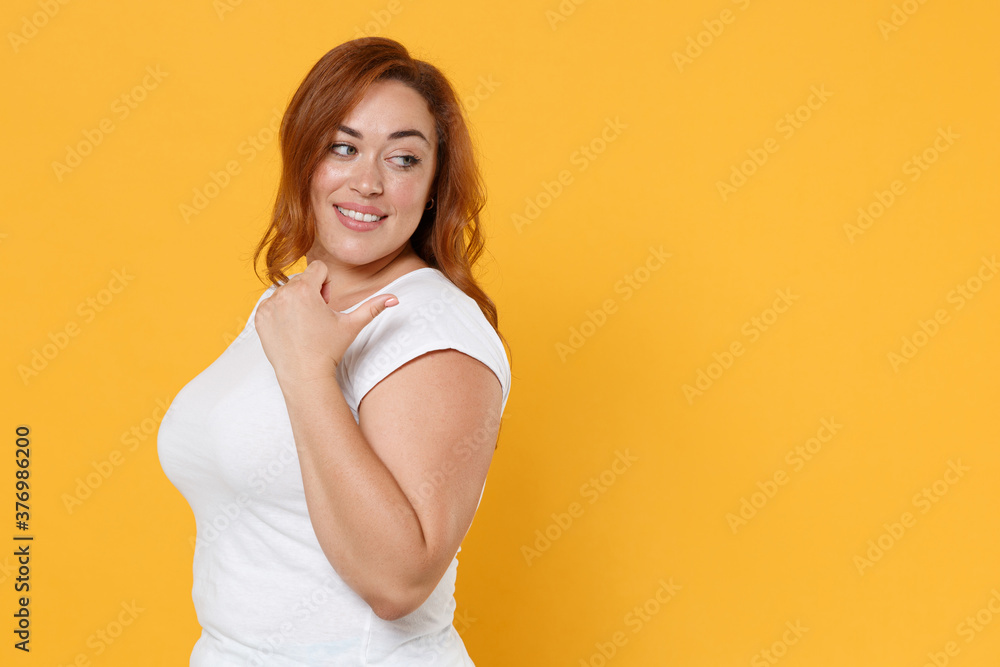 Side view of smiling young redhead plus size body positive female woman in white casual t-shirt posing pointing thumb aside on mock up copy space isolated on yellow color background studio portrait. Side view of smiling young redhead plus size body positive female woman in white casual t-shirt posing pointing thumb aside on mock up copy space isolated on yellow color background studio portrait.