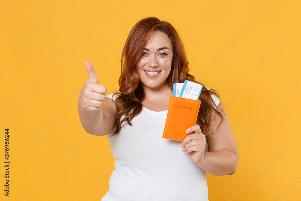 Smiling young redhead plus size body positive traveler tourist female woman girl 20s in white casual t-shirt hold passport tickets showing thumb up isolated on yellow color background studio portrait. Smiling young redhead plus size body positive traveler tourist female woman girl 20s in white casual t-shirt hold passport tickets showing thumb up isolated on yellow color background studio portrait.