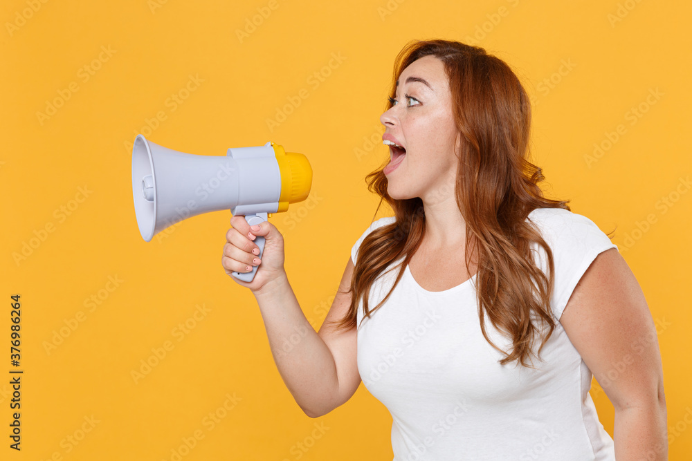 Shocked excited young redhead plus size body positive female woman girl 20s in white casual t-shirt posing screaming in megaphone looking aside isolated on yellow color background studio portrait. Shocked excited young redhead plus size body positive female woman girl 20s in white casual t-shirt posing screaming in megaphone looking aside isolated on yellow color background studio portrait.