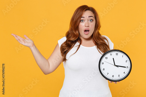 Shocked worried young redhead plus size body positive female woman girl in white casual t-shirt posing holding clock spreading hands looking camera isolated on yellow color background studio portrait.