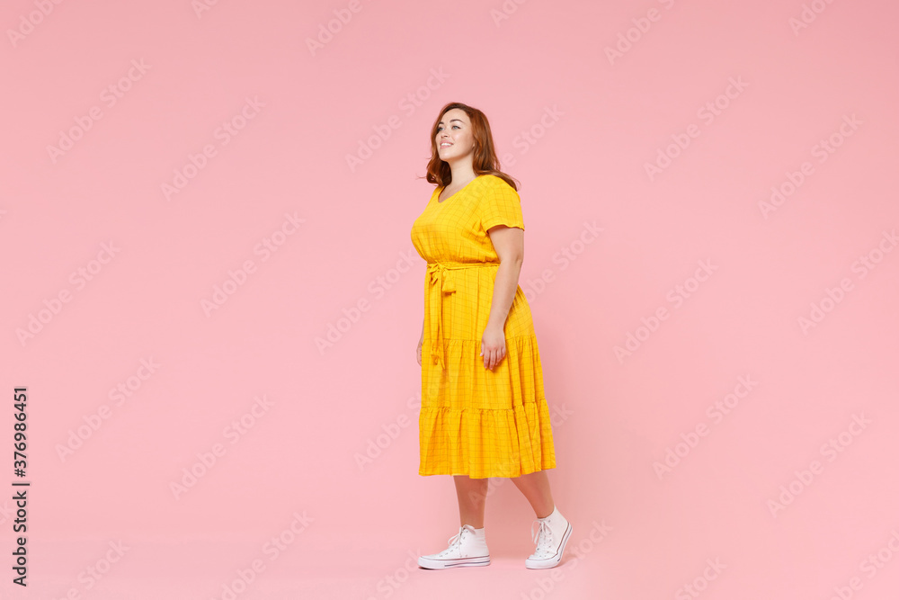 Full length portrait of smiling attractive beautiful young redhead plus size body positive female woman girl 20s in yellow dress posing looking aside isolated on pastel pink color background studio. Full length portrait of smiling attractive beautiful young redhead plus size body positive female woman girl 20s in yellow dress posing looking aside isolated on pastel pink color background studio.
