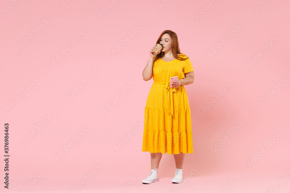 Full length portrait pretty young redhead plus size body positive female woman 20s in yellow dress hold paper cup coffee tea mobile phone looking aside isolated on pastel pink color background studio. Full length portrait pretty young redhead plus size body positive female woman 20s in yellow dress hold paper cup coffee tea mobile phone looking aside isolated on pastel pink color background studio.