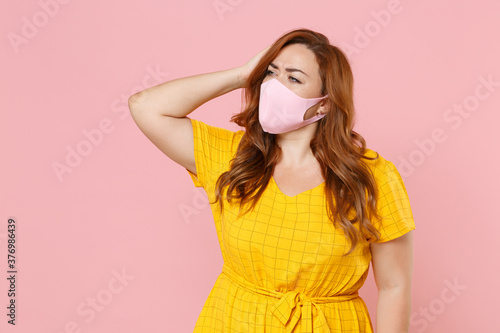Preoccupied young plus size body positive female woman in yellow dress face mask to safe from coronavirus virus covid-19 put hand on head looking aside isolated on pink background studio portrait.