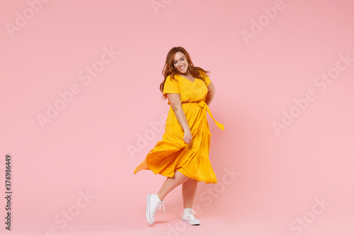 Full length portrait of smiling beautiful charming young redhead plus size body positive female woman girl 20s in yellow dress posing looking camera isolated on pastel pink color background studio.