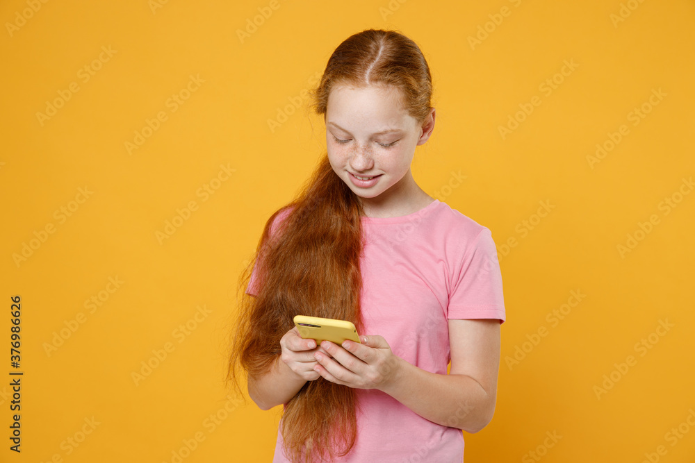 Smiling little ginger redhead kid girl 12-13 years old in pink casual t-shirt posing using mobile cell phone typing sms message isolated on bright yellow color background children studio portrait. Smiling little ginger redhead kid girl 12-13 years old in pink casual t-shirt posing using mobile cell phone typing sms message isolated on bright yellow color background children studio portrait.