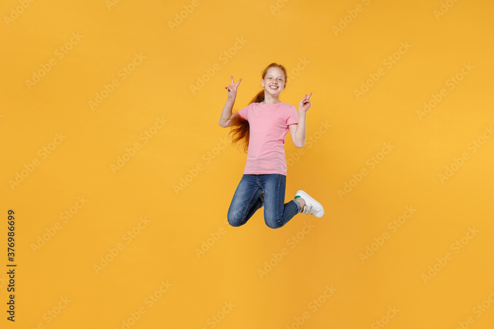 Full length children studio portrait of smiling little ginger redhead kid girl 12-13 years old in pink casual t-shirt posing jumping showing victory sign isolated on bright yellow color background.