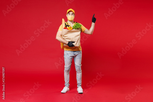 Full length delivery employee man in yellow cap face mask gloves t-shirt thermal backpack hold paper takeaway food bag products work courier service in quarantine covid-19 isolated on red background.