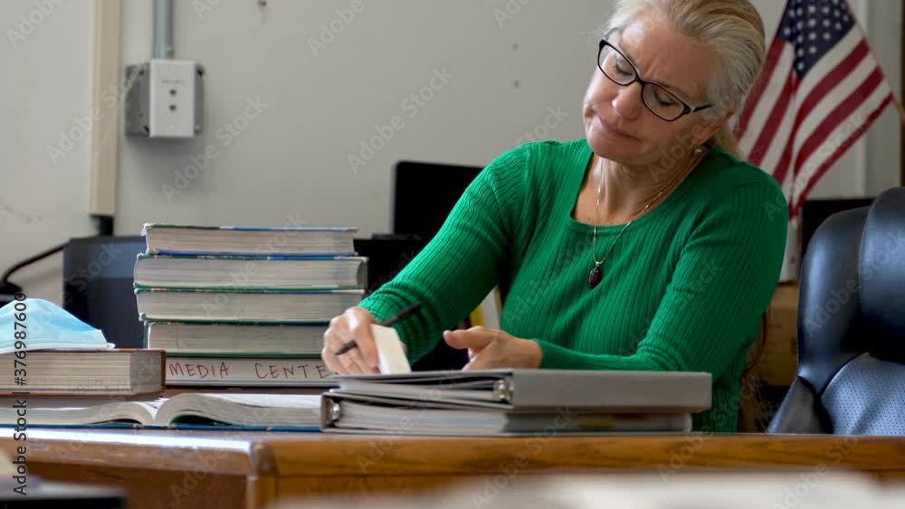 Closeup of teacher in school working at desk with wearing medical face mask and sanitizer on desk with PPE and US flag behind. Closeup of teacher in school working at desk with wearing medical face mask and sanitizer on desk with PPE and US flag behind.