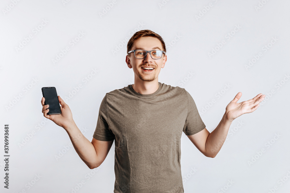 Young handsome man holding a smartphone and smiling. The guy shows his hands and raises his shoulders. Student, worker posing for photo isolated on gray background. Place for advertising. Young handsome man holding a smartphone and smiling. The guy shows his hands and raises his shoulders. Student, worker posing for photo isolated on gray background. Place for advertising.