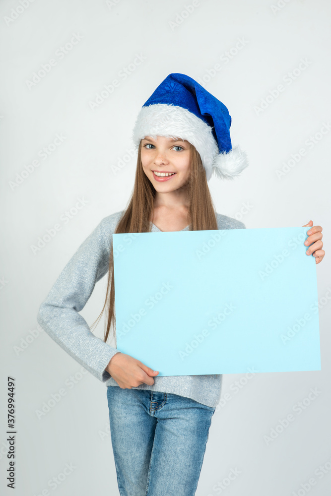Cute little girl in winter clothes with blue Santa hat posing on light background with blank paper nameplate in hand. Cute little girl in winter clothes with blue Santa hat posing on light background with blank paper nameplate in hand.