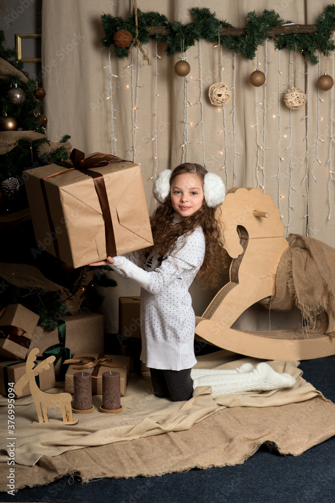 Cute little girl in winter clothes posing in home front of Christmas tree with gift box in hand.