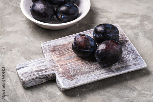 Fresh ripe plum fruits with water drops in wooden bowl and cutting board on stone concrete background, angle view