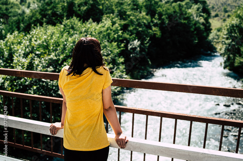 Unrecognizable young woman stands on bridge and enjoys beautiful view of mountain river which flowing between green trees. Rear view of female tourist looking at clean waterway