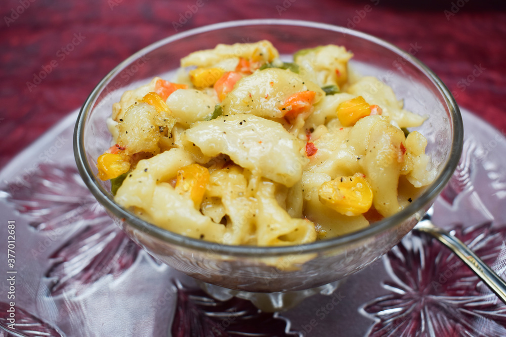 Close up of Vegetable macaroni cooked in White sauce served in a transparent bowl isolated on textured background. Cooked pasta with carrot, corn, capsicum sprinkled oregano and chillyflakes Close up of Vegetable macaroni cooked in White sauce served in a transparent bowl isolated on textured background. Cooked pasta with carrot, corn, capsicum sprinkled oregano and chillyflakes