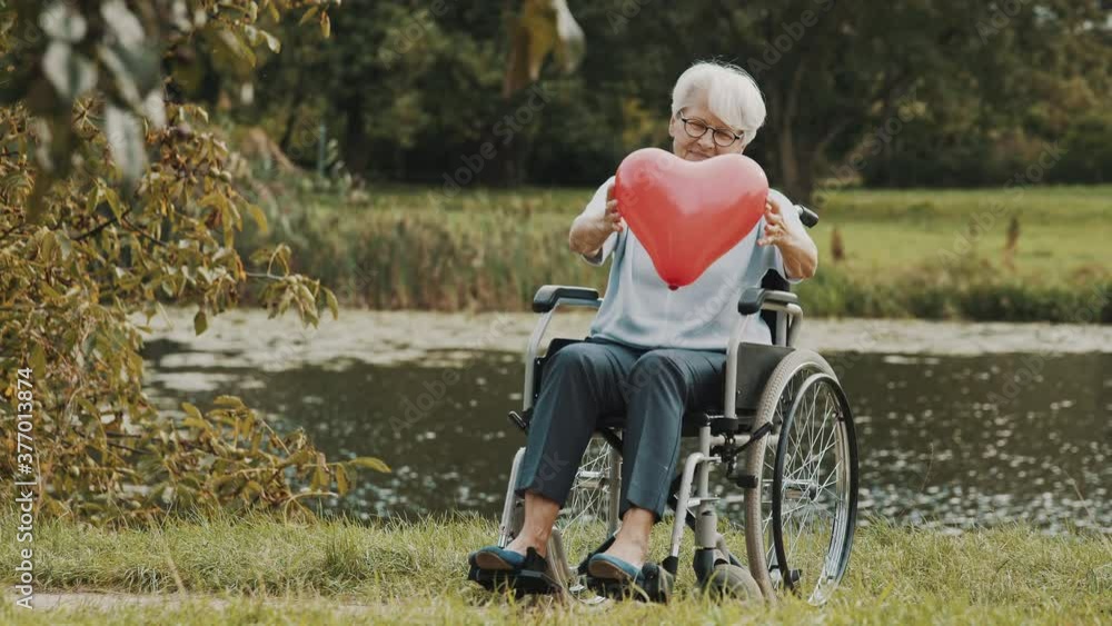 senior woman in the wheelchair near the river holding heart shaped balloon. High quality 4k footage senior woman in the wheelchair near the river holding heart shaped balloon. High quality 4k footage