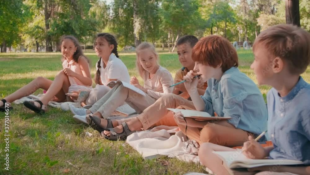 Slowmo medium shot of happy schoolchildren with notebooks sitting on blankets in park and laughing while talking to teacher off camera Slowmo medium shot of happy schoolchildren with notebooks sitting on blankets in park and laughing while talking to teacher off camera