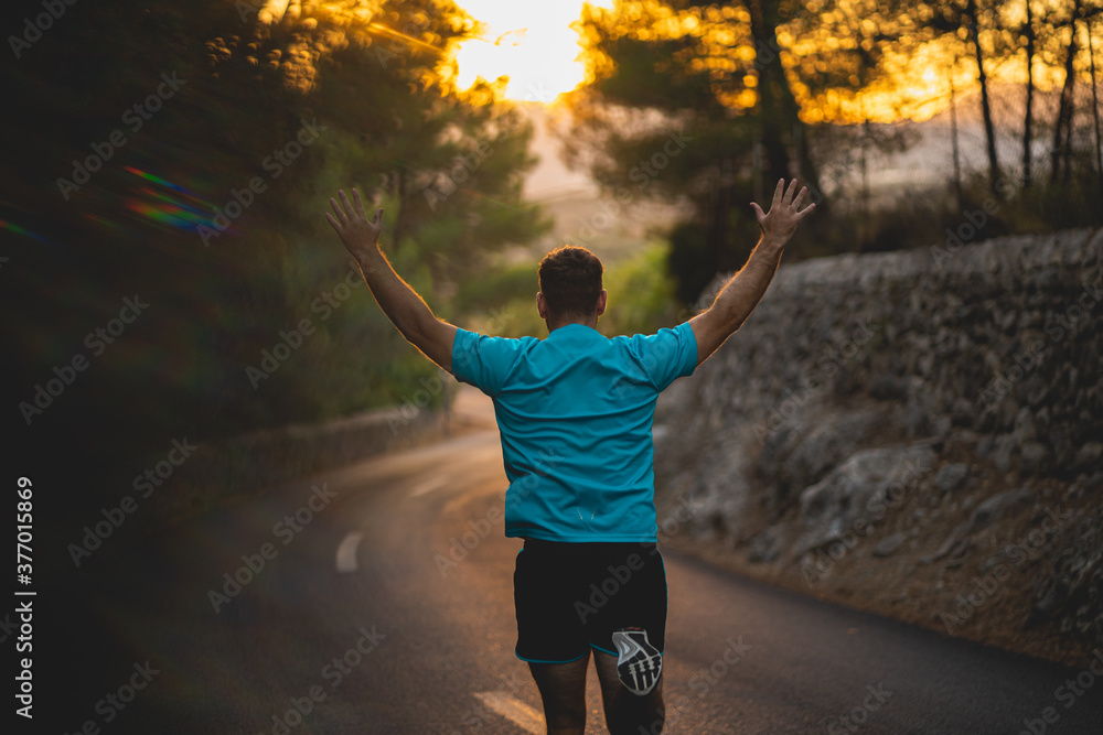 Young man wearing blue shirt and black shorts runs while enjoying freedom on an idyllic road during sunset surrounded by nature in Mallorca (Spain)