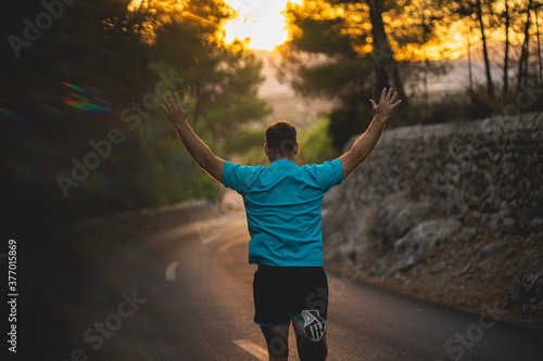 Young man wearing blue shirt and black shorts runs while enjoying freedom on an idyllic road during sunset surrounded by nature in Mallorca (Spain)