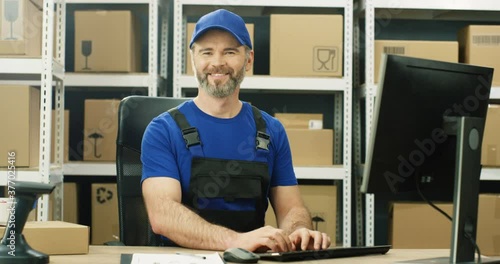 Portrait shot of cheerful Caucasian postman in iniform and cap sitting at table in postal office store and working at computer. Handsome male courier smiling happily to camera in storage of parcels. photo