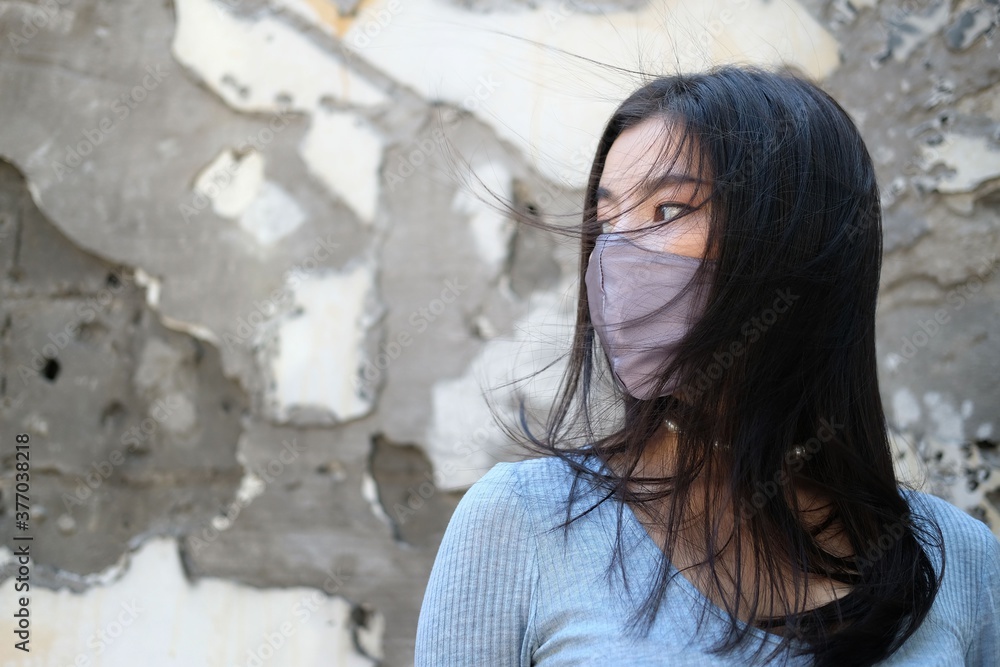 portrait of of asian girl in protective sterile mask on her face looking at camera outdoors  with copy space of half turned attractive on a wall decay background