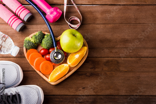 Top view of fresh fruits and vegetables in heart plate wood (apple, carrot, tomato, orange, broccoli) and sports equipment and doctor stethoscope on wooden table, Healthy lifestyle diet food concept