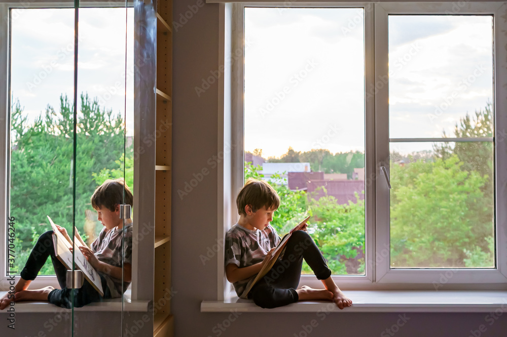 The boy reads a book while sitting on the windowsill. His reflection is visible on the mirror of the cabinet