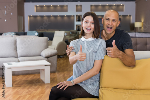 Portrait of happy couple with thumbs up sitting on yellow sofa in furniture shop
