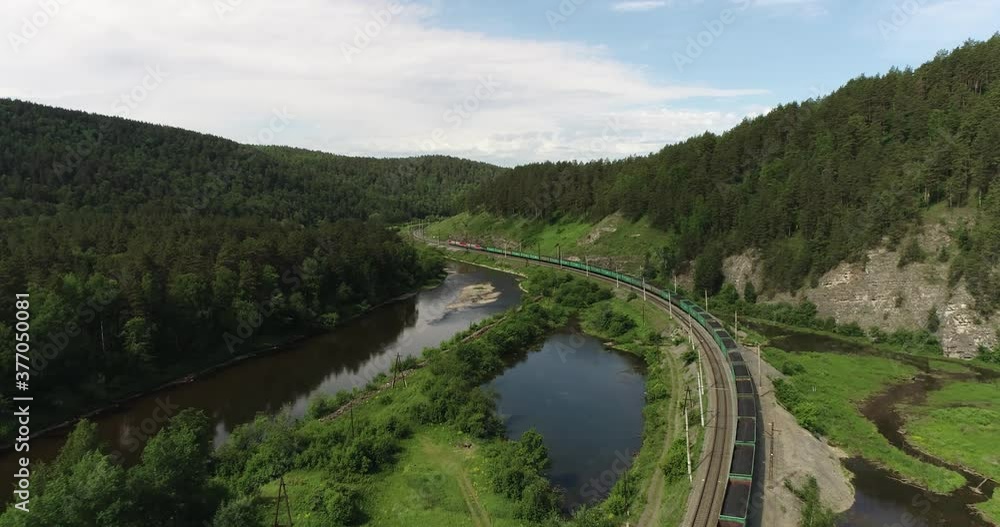 Freight long train carries with coal carriages an electric locomotive by two-sided Trans Siberian railways under the rock and near mountains river / Aerial drone wide view at summer sunny day Freight long train carries with coal carriages an electric locomotive by two-sided Trans Siberian railways under the rock and near mountains river / Aerial drone wide view at summer sunny day