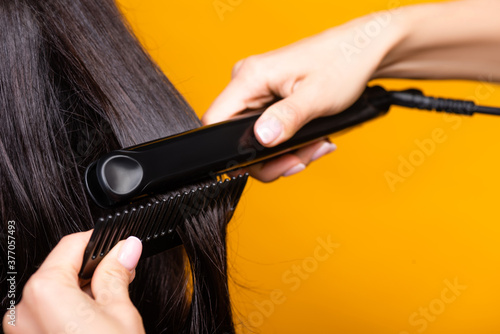 cropped view of hairdresser using hair straightener and comb isolated on yellow