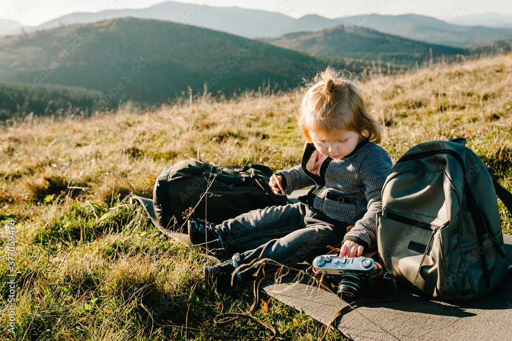 Little hiker girl with camera and backpack sitting on grass in autumn mountains. Travel concept. Holiday, trip and vacation. World Tourism Day.