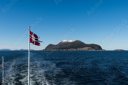Norwegian mail or post flag hoisted on post of mailboat waving in the wind with open sea and mountains on sunny winter day