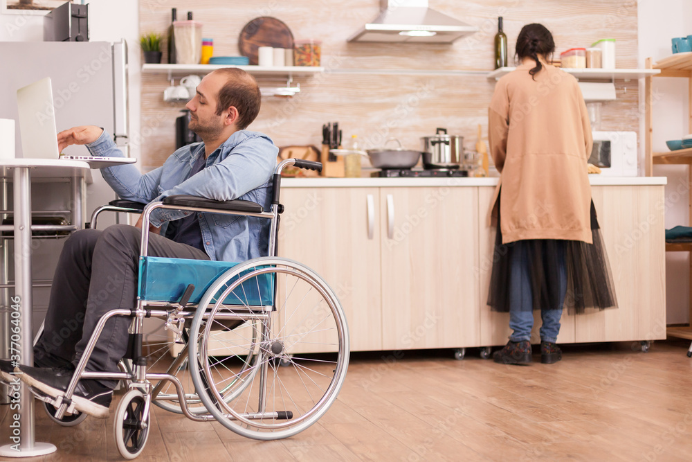 Guy with walking disability in wheelchair using laptop in kitchen and wife is cooking meal. Disabled paralyzed handicapped man with walking disability integrating after an accident.