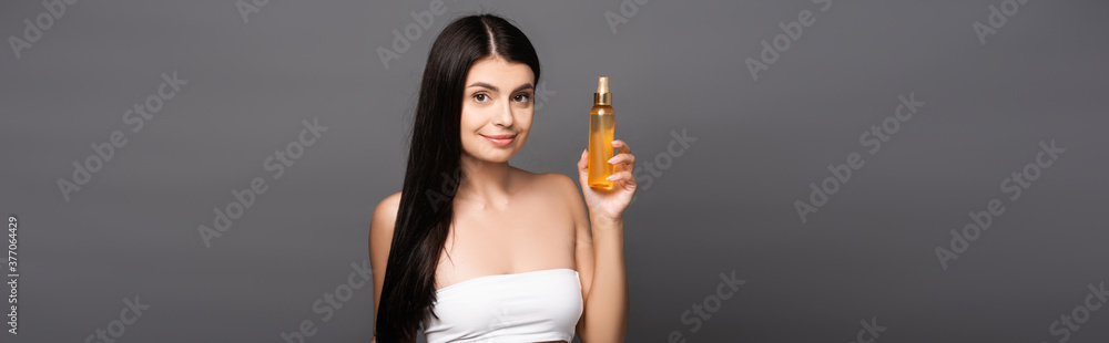 brunette woman holding hair oil in spray bottle and smiling isolated on black, panoramic shot brunette woman holding hair oil in spray bottle and smiling isolated on black, panoramic shot