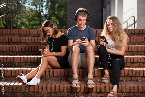 Digital technology addiction concept. Three young students sitting on the stairs and concentrated on their smartphones.