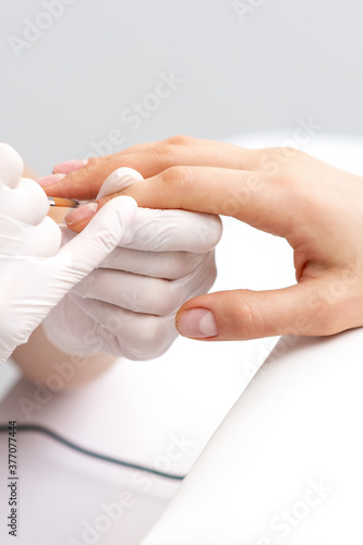 Manicure master in rubber gloves applying transparent nail polish on female nails in beauty salon