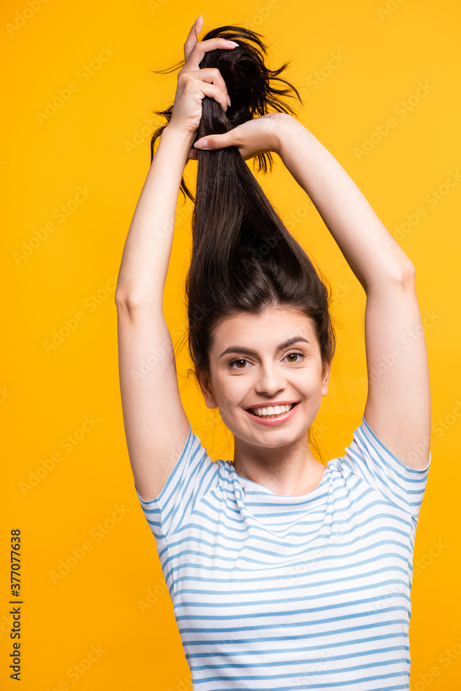 Fototapeta premium brunette woman holding hair above head and smiling isolated on yellow