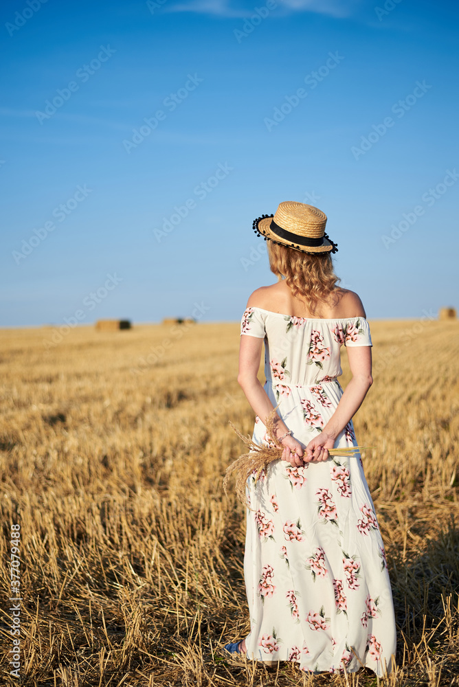 Young blond woman, wearing white romantic dress and straw hat, with back to camera, holding dried grass bouquet standing on straw field in summer. Female portrait on natural background.