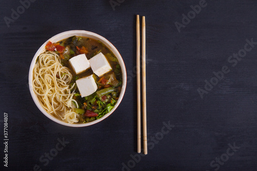 Japanese soup with tofu, noodles, vegetables and herbs, bamboo chopsticks on a black background. Copy spaes.