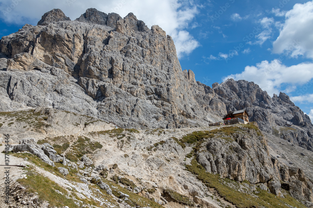 Mountain hut stands below an imposing mountain peak at the end of a steep trail. Catinaccio - Dolomites, Italy