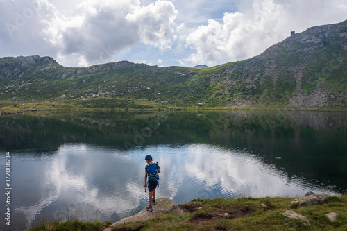Kid standing on a rock on the edge of the Alpine Bombasel Lake on Cermis Alps - Italy. Dolomite peaks are visible in the background and reflect in the water along with the clouds. Trentino, Italy © Travelvolo