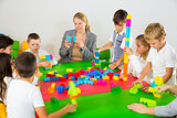 Group of school kids with teacher sitting together around desk in classroom, playing with plastic building blocks