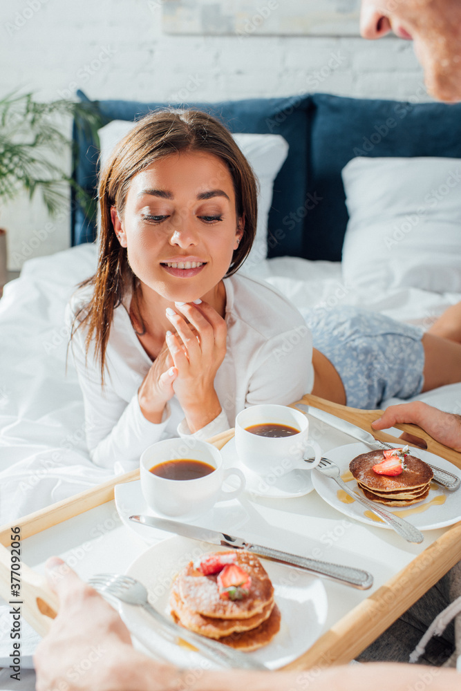 Selective focus of man holding pancakes and coffee near woman on bed