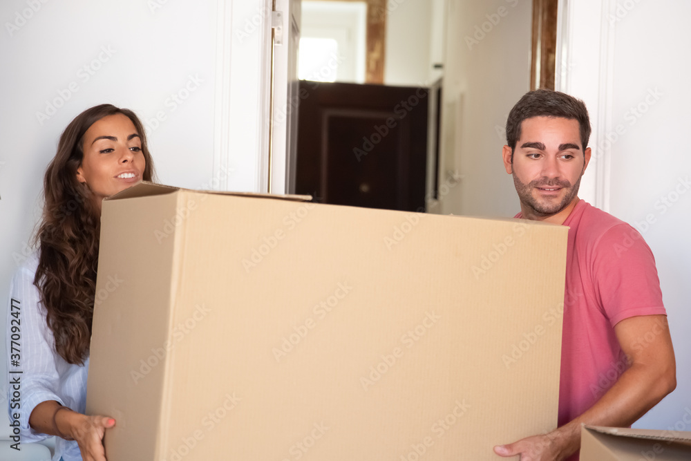 Positive young family couple moving into new house, carrying carton box. Medium shot. Real estate or property buying concept Positive young family couple moving into new house, carrying carton box. Medium shot. Real estate or property buying concept